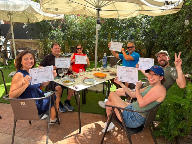 Workshop guests at a table under the umbrellas holding up their paella certificates